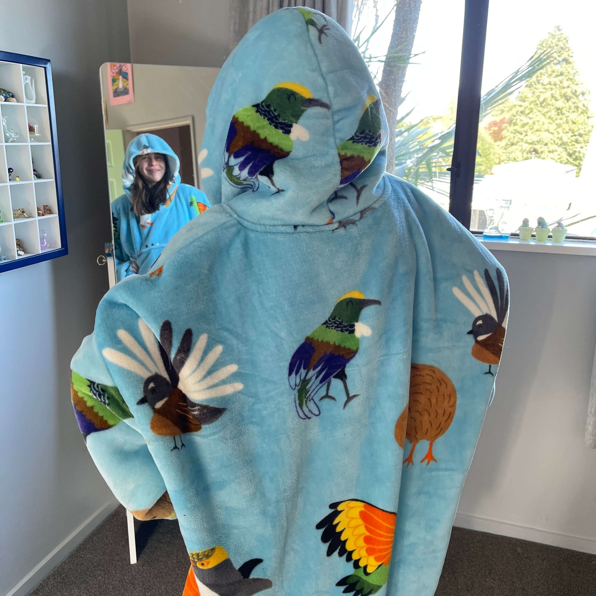 Girl looking at herself in a mirror wearing an oversized blue hoodie with New Zealand birds printed on it.