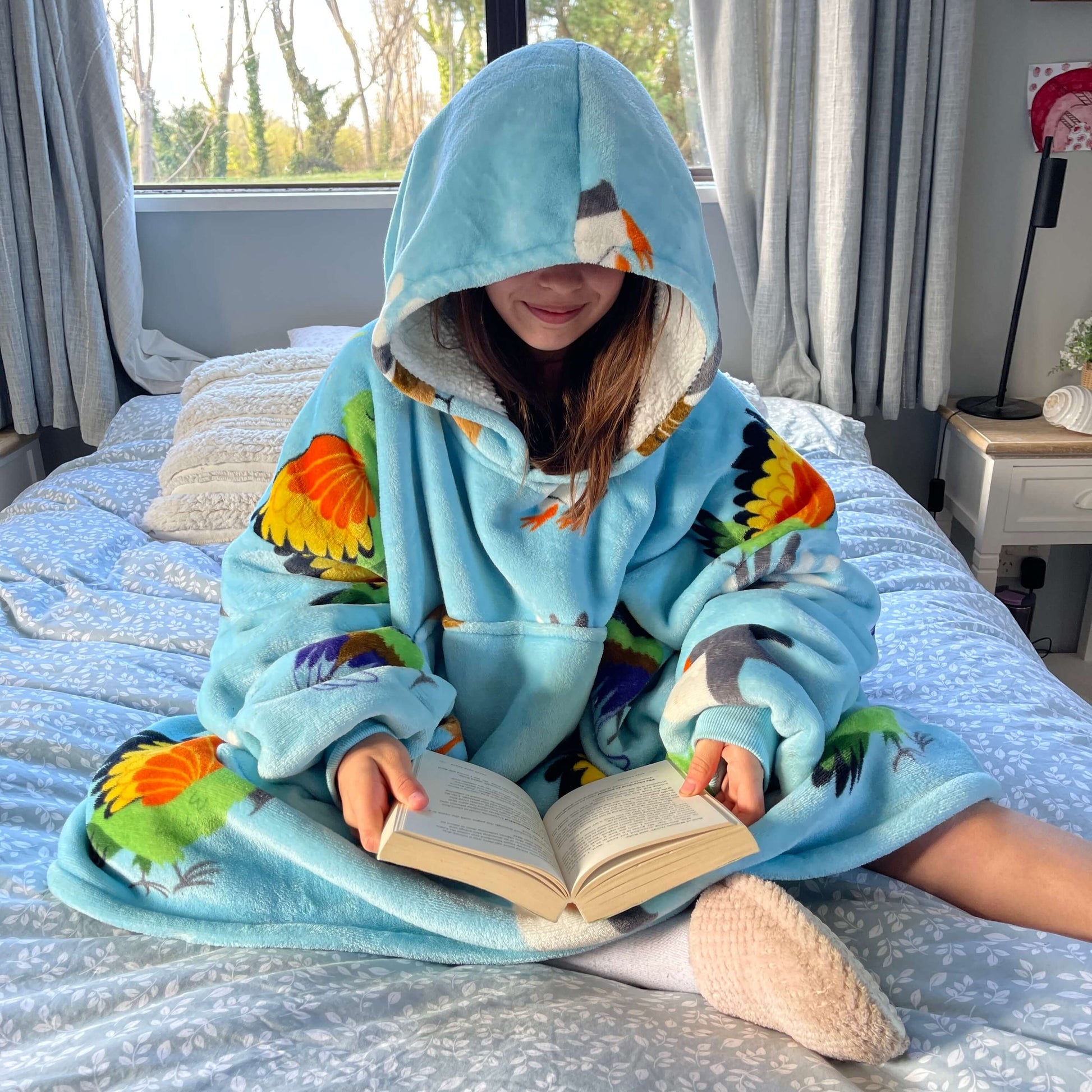 Girl sitting on a bed reading a book wearing an oversized blue hoodie with New Zealand birds printed on it.
