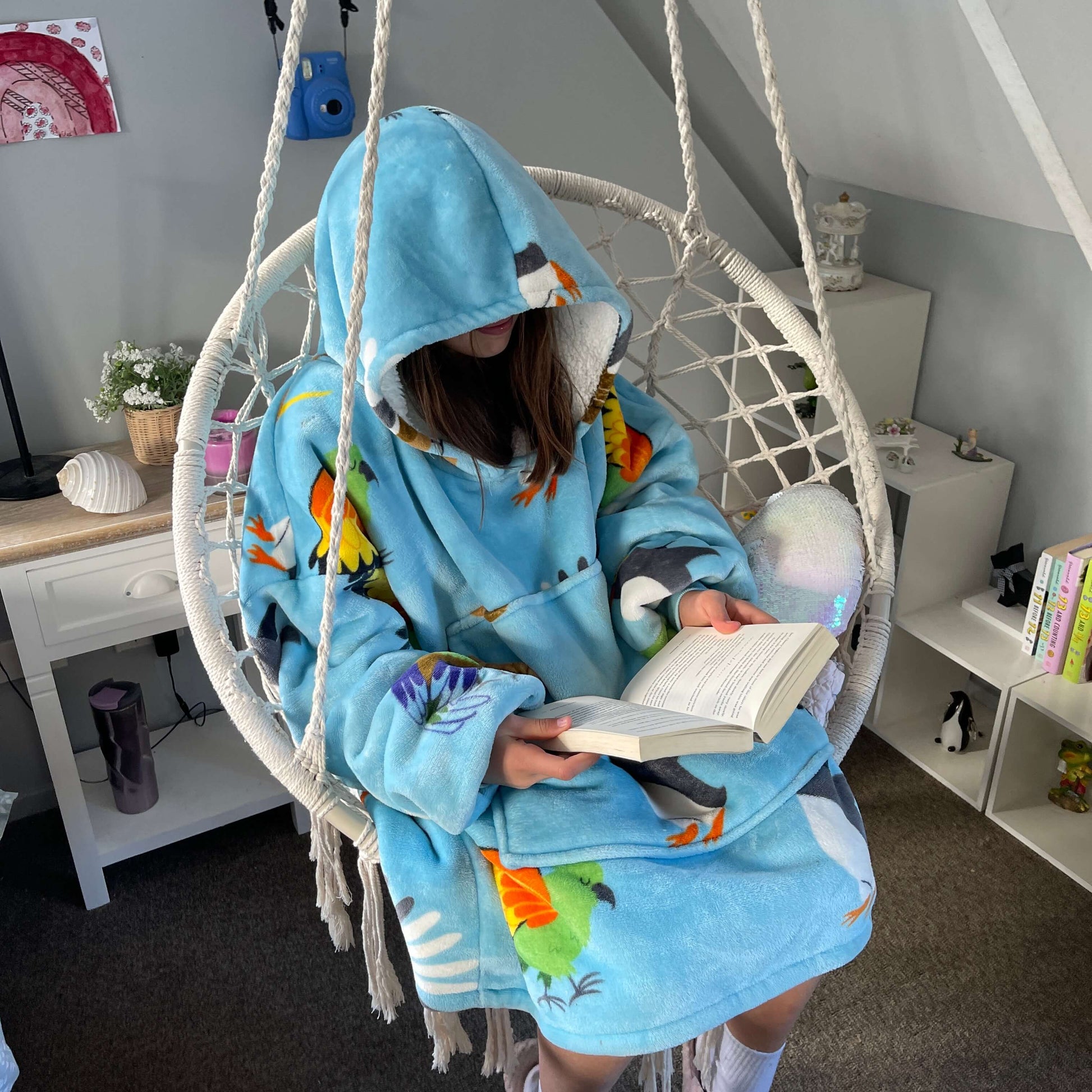 Girl sitting in a swing chair reading a book wearing an oversized blue hoodie with New Zealand birds printed on it.