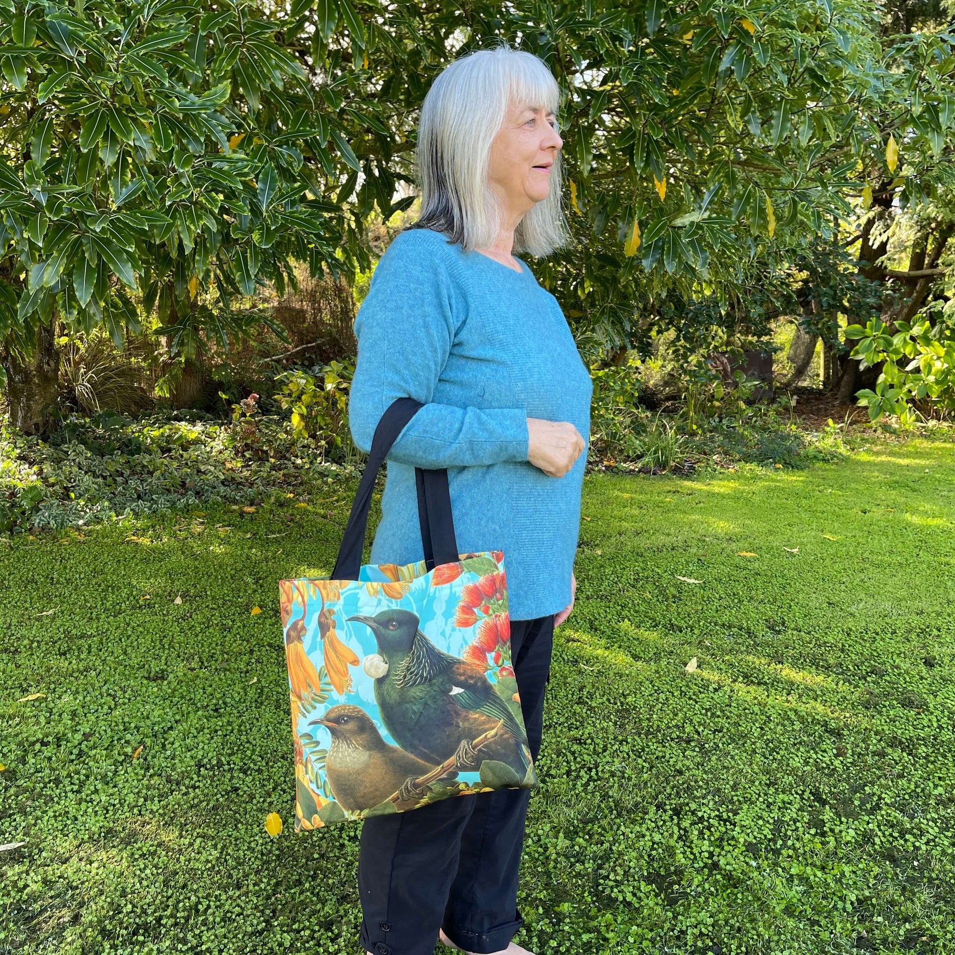 Woman with a tote bag over her arm. The tote features a Bellbird & Tui amongst Kowhai & Pohutukawa flowers.