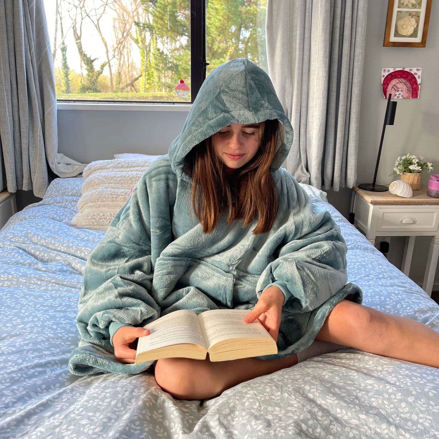 Girl sitting on a bed reading a book wearing an oversized blue hoodie.