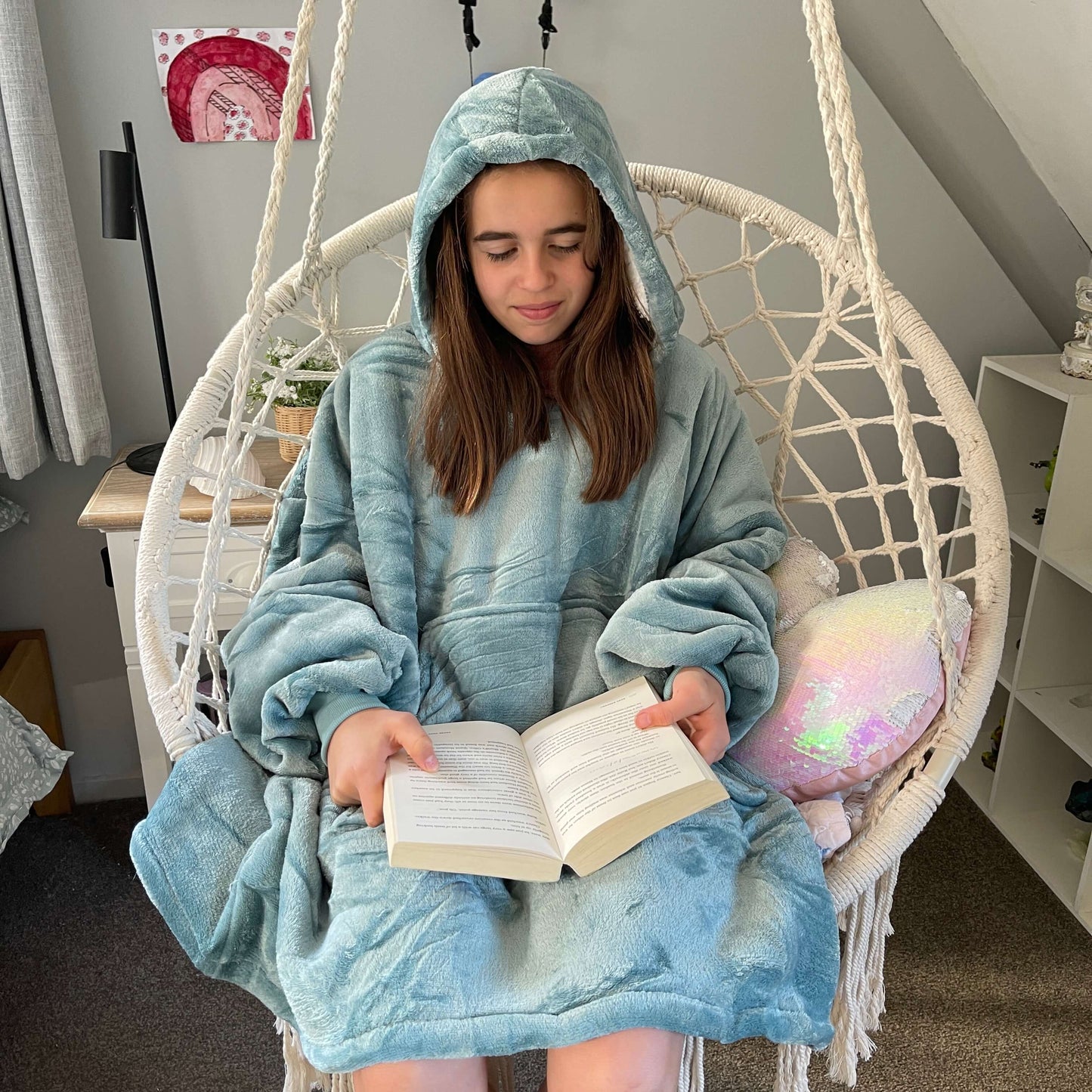 Girl sitting in a swing chair reading a book wearing an oversized blue hoodie.