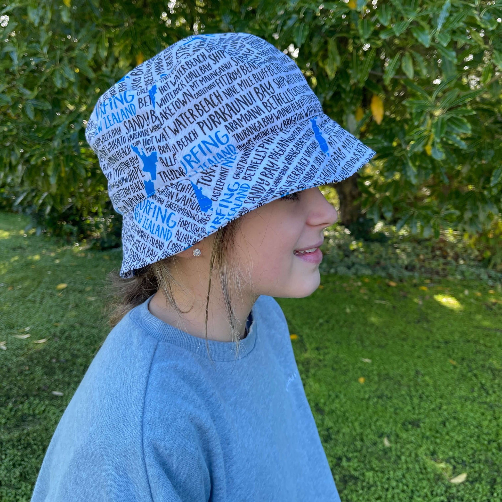 Girl wearing a bucket hat in white with blue and black text stating New Zealand surfing spots.