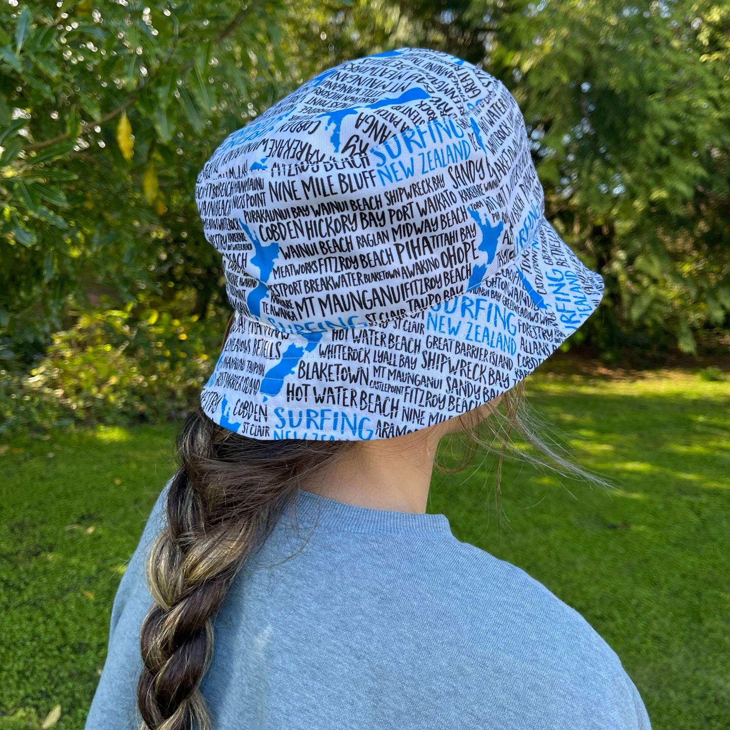 Girl wearing a bucket hat in white with blue and black text stating New Zealand surfing spots.