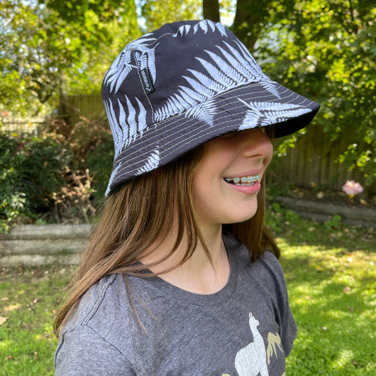 Side profile of a young girl with long brown hair head wearing a bucket hat in black with white ferns printed on it.
