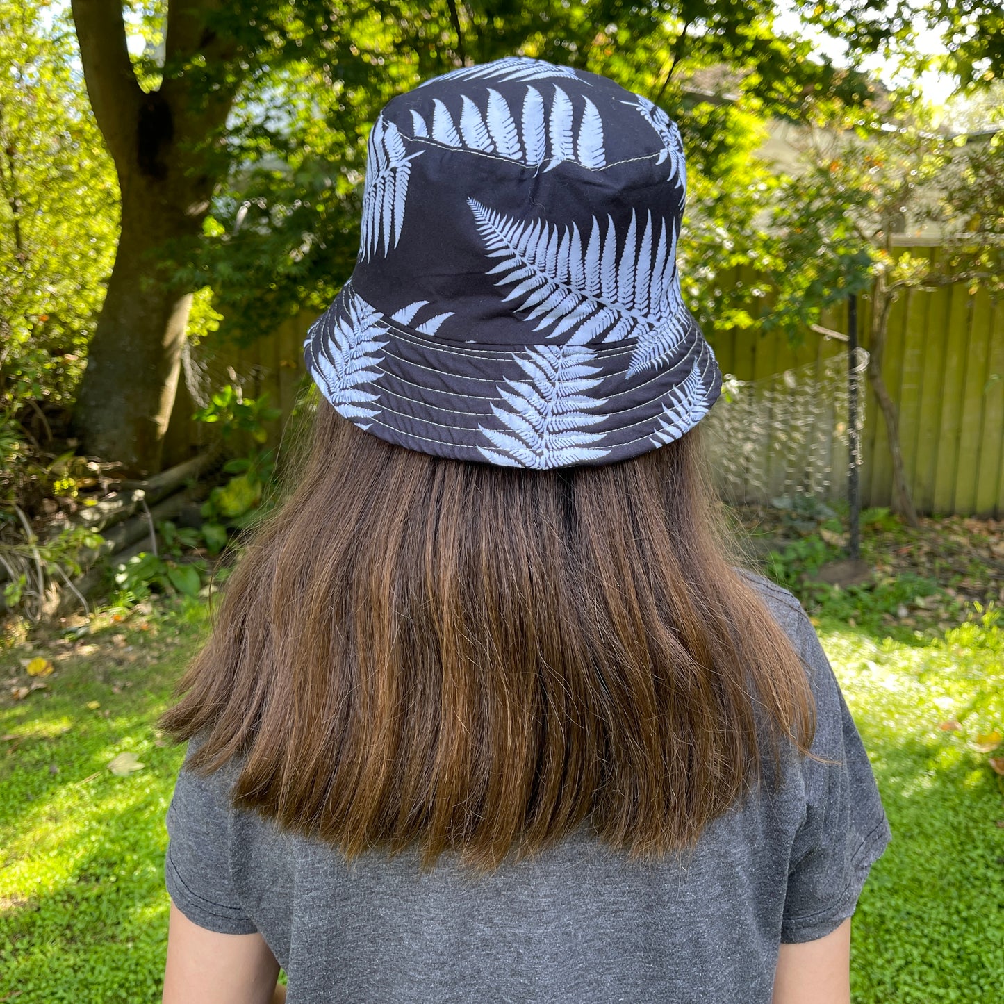 Back of a young girl with long brown hair head wearing a bucket hat in black with white ferns printed on it.
