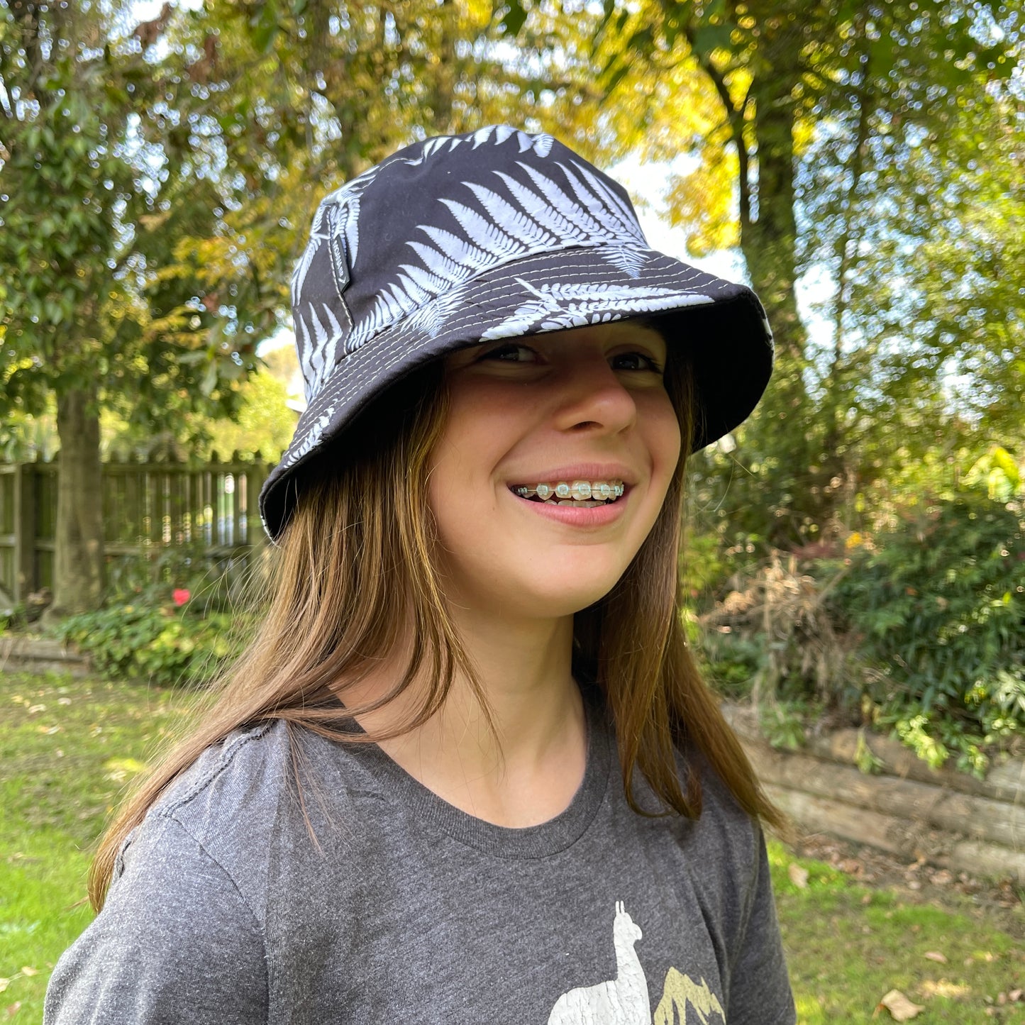 Young girl with long brown hair head wearing a bucket hat in black with white ferns printed on it.