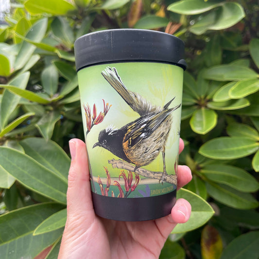 Black reusable coffee cup with Hihi birds and Flax flowers on a green background.