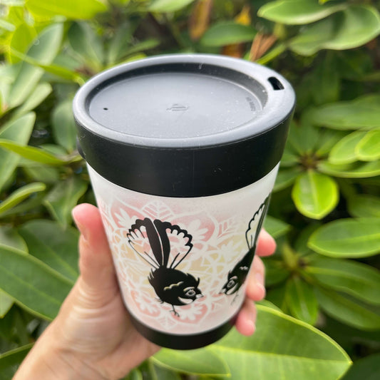 Black reusable coffee cup with white wrap and yellow & pink floral design featuring two cartoon style Fantail birds, showcasing the lid with sipper hole.