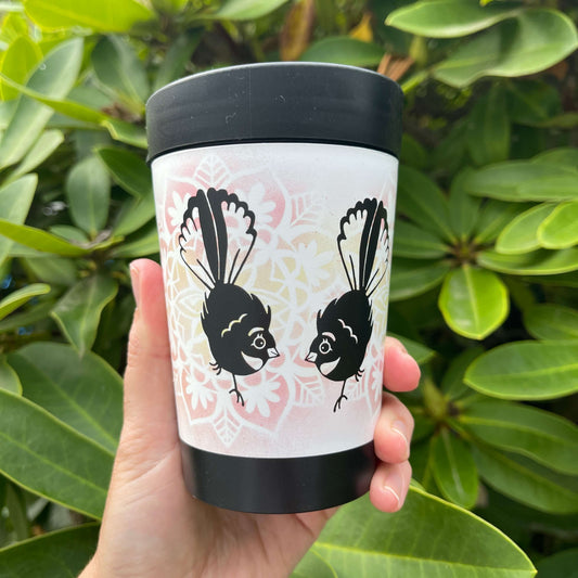 Black reusable coffee cup with white wrap and yellow & pink floral design featuring two cartoon style Fantail birds.