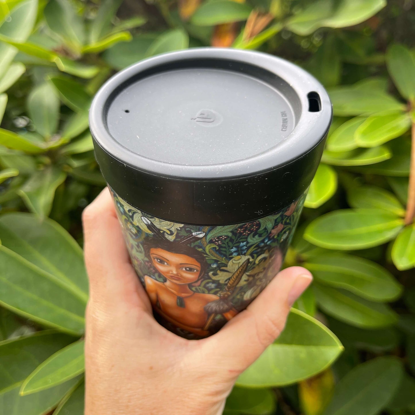 Black reusable coffee cup featuring a floral wrap and featuring an artists image of a young Māori boy with most of the image showing the black lid and sipper hole.