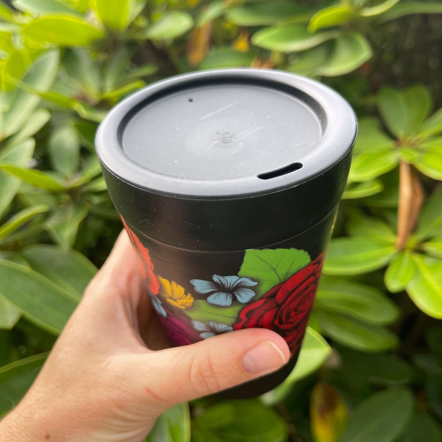 Black reusable coffee cup with bright bold flowers wrapped around it showing the lid with sipper hole.