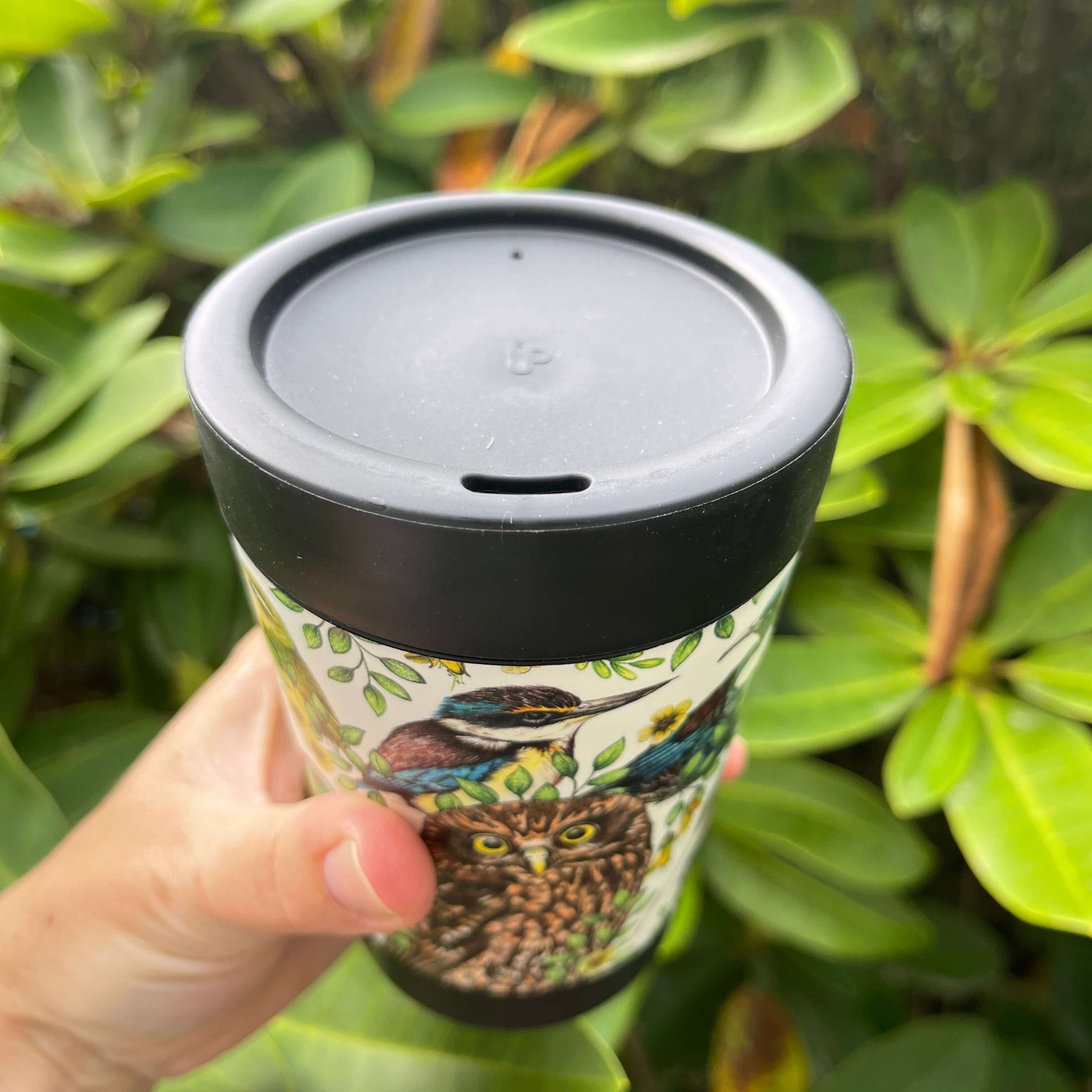 Black reusable coffee cup wrapped with beautiful drawings of New Zealand birds showing the lid with the sipper hole.