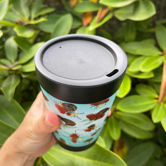 Black reusable coffee cup with blue ombre wrap featuring Kiwi birds and bunches of Pohutukawa flowers, Kowhai flowers and Ferns, showing the lid with sipper hole.