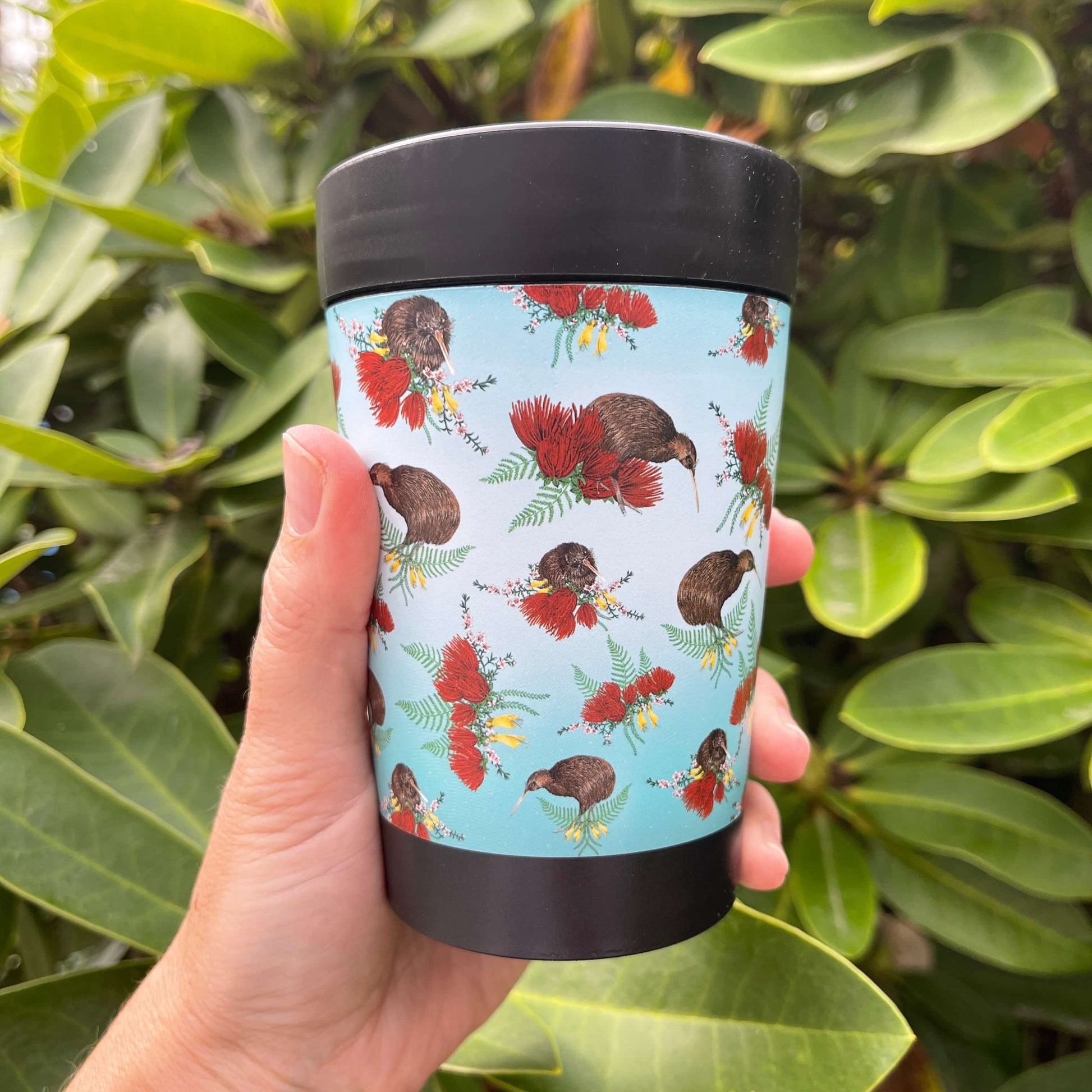 Black reusable coffee cup with blue ombre wrap featuring Kiwi birds and bunches of Pohutukawa flowers, Kowhai flowers and Ferns.