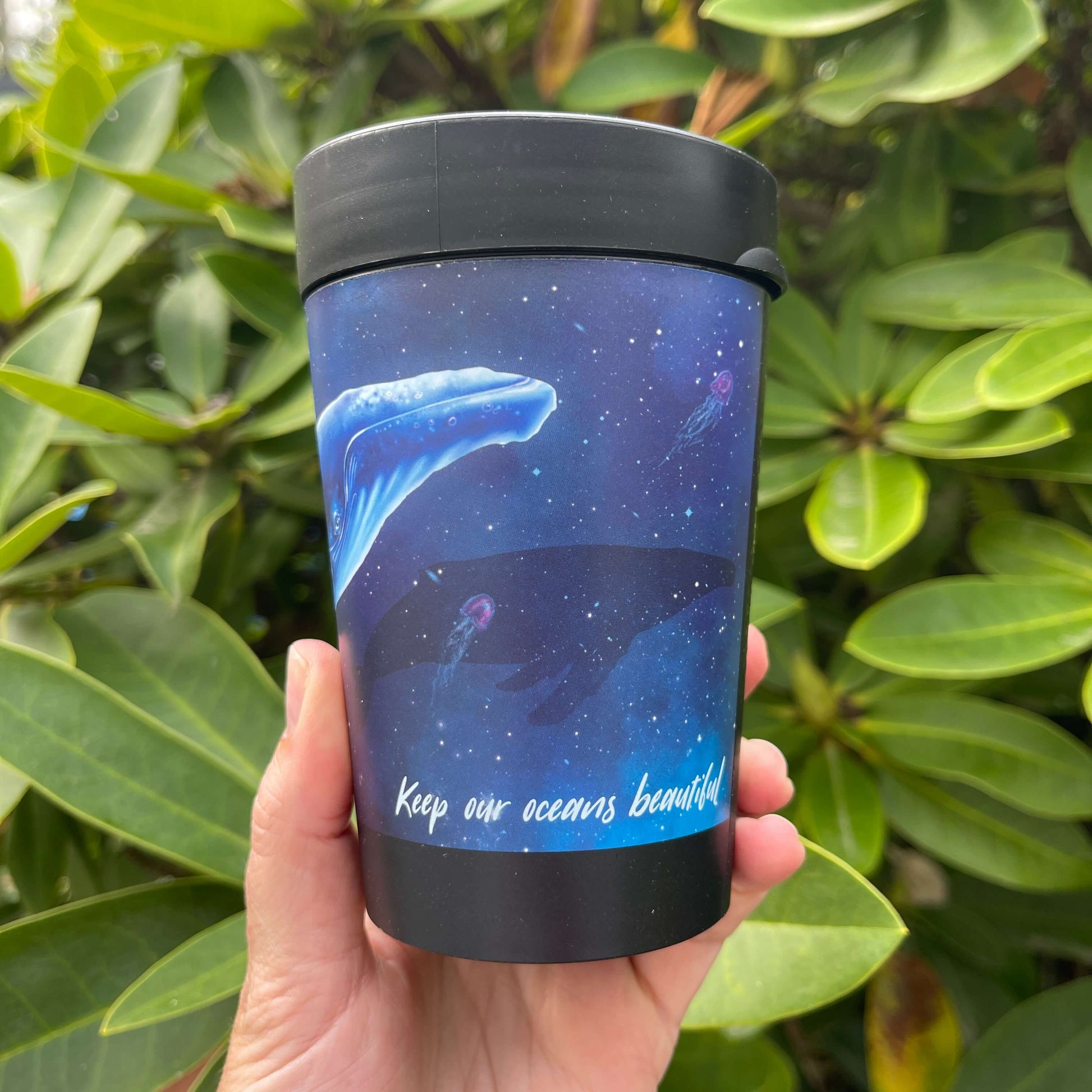 Black reusable coffee cup with blue galaxy wrap featuring a Humpback whale and the words "Keep our oceans beautiful".