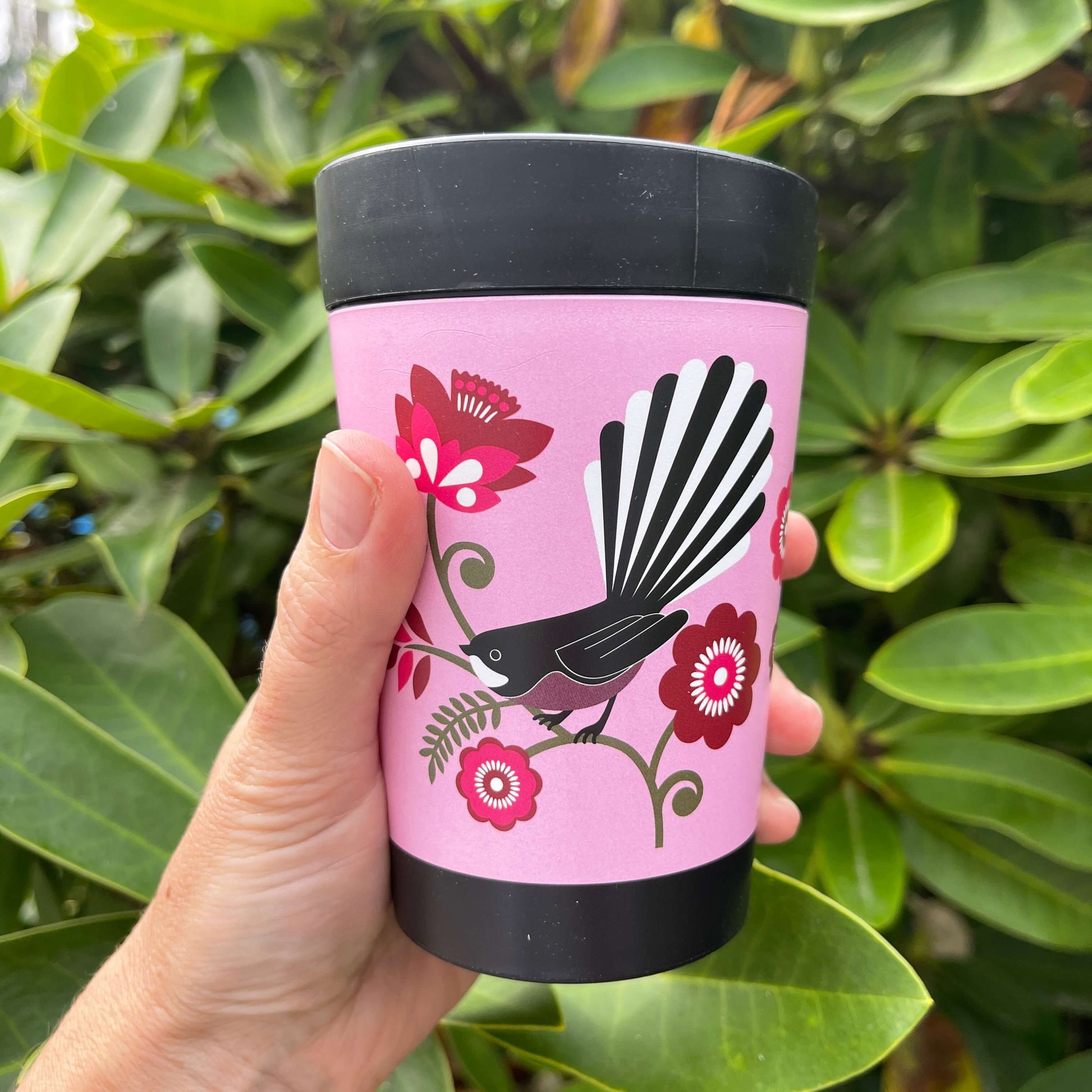 Black reusable coffee cup with bright pink wrap featuring a Fantail bird sitting on a green koru branch with pink flowers.