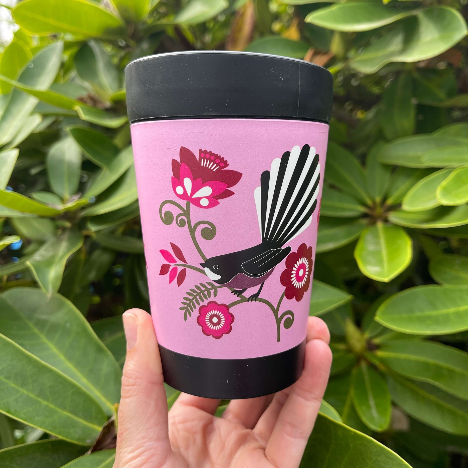 Black reusable coffee cup with bright pink wrap featuring a Fantail bird sitting on a green koru branch with pink flowers.