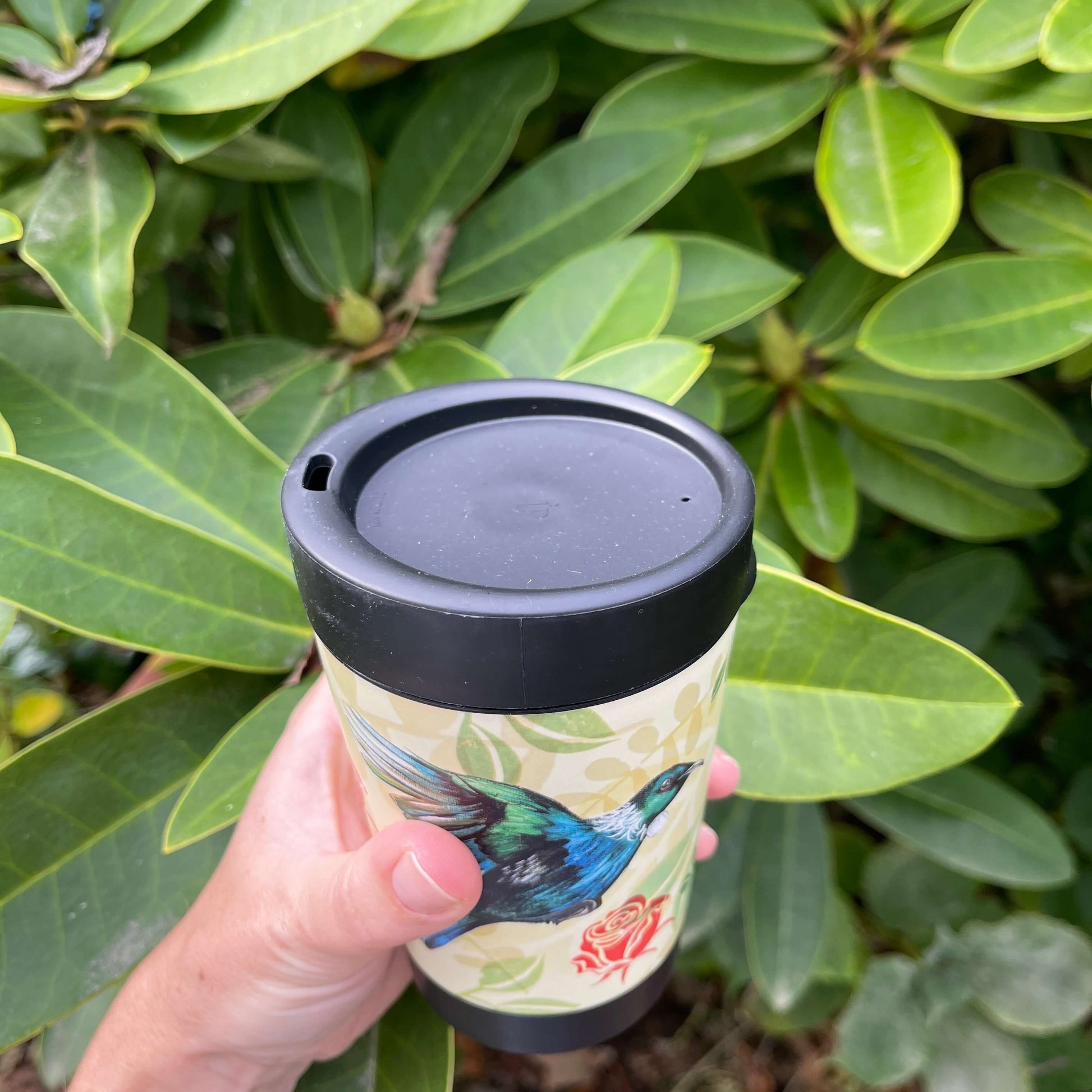 Black reusable coffee cup with beige and green leaf patterned wrap featuring the New Zealand Tui bird and red roses. Showing the lid with a sipper hole.