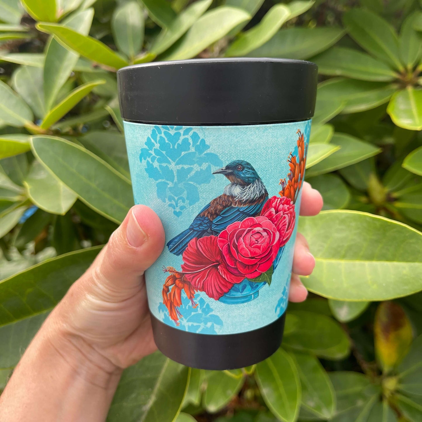 Black reusable coffee cup with light blue patterned wrap featuring a Tūī bird on a branch of pink and orange flowers.