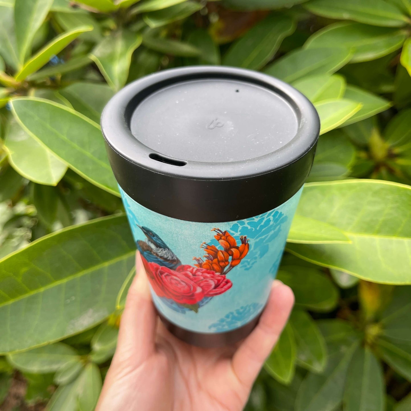 Black reusable coffee cup with light blue patterned wrap featuring a Tūī bird on a branch of pink and orange flowers, highlighting the lid with has a sipper hole.