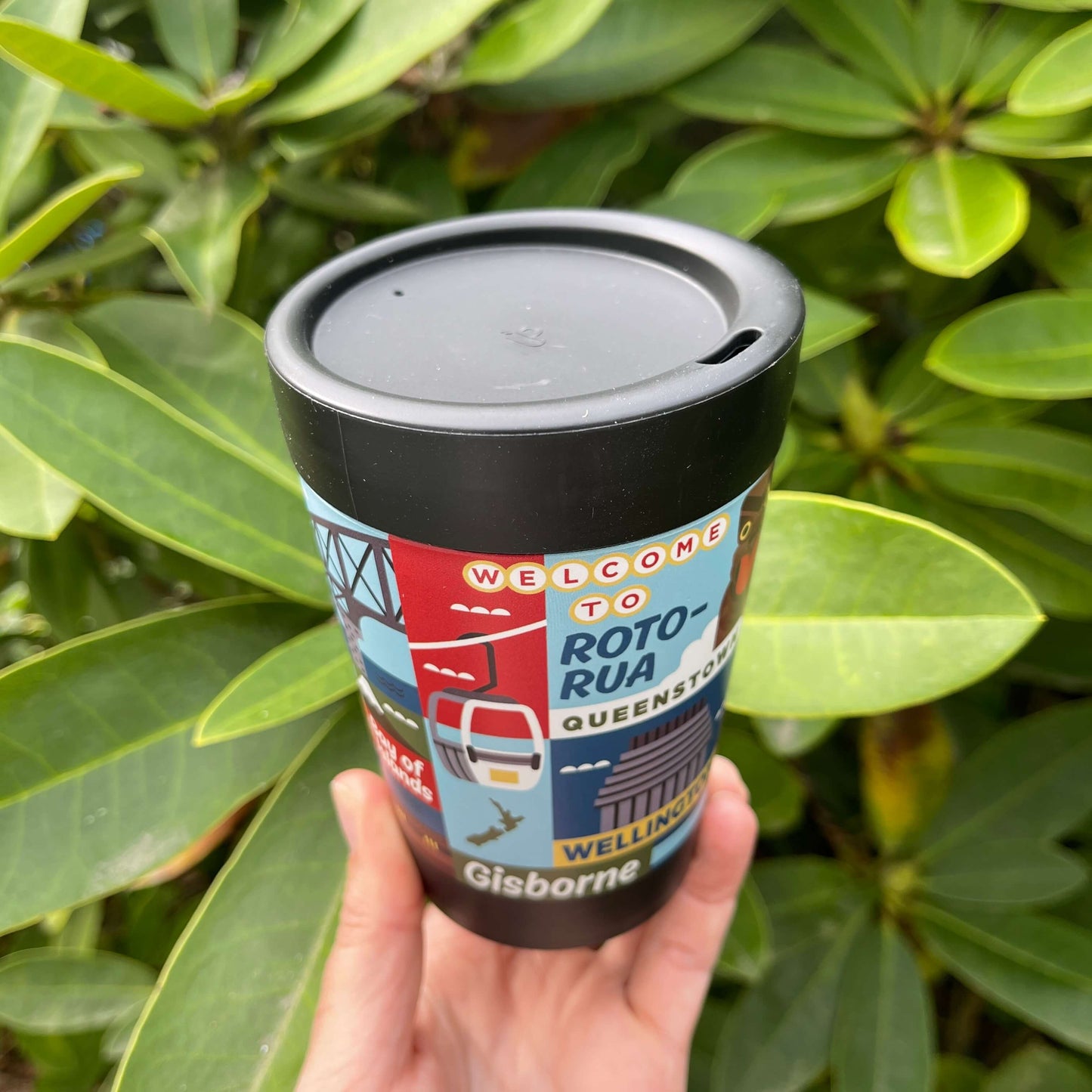 Reusable coffee cup in black with artist images of New Zealand towns wrapped around it and showing the black lid with the sipping hole.