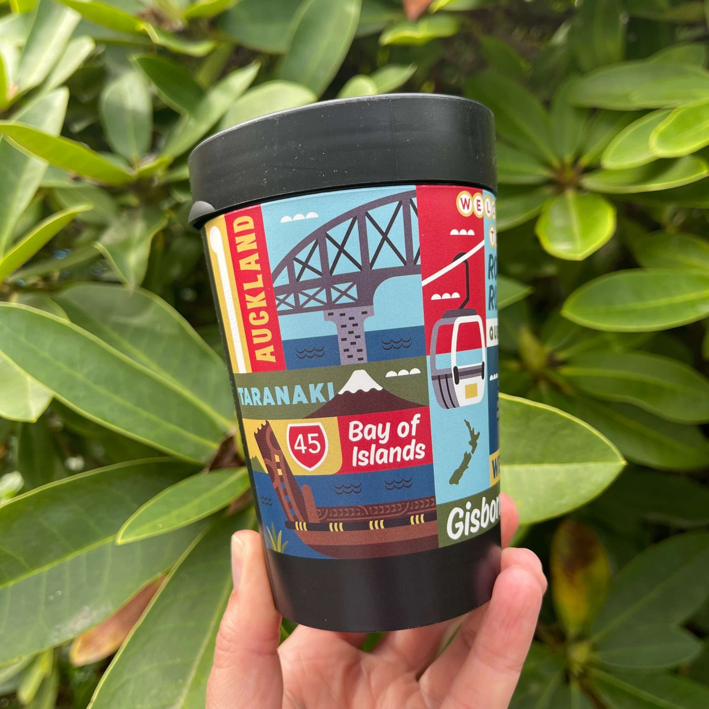 Reusable coffee cup in black with artist images of New Zealand towns wrapped around it.