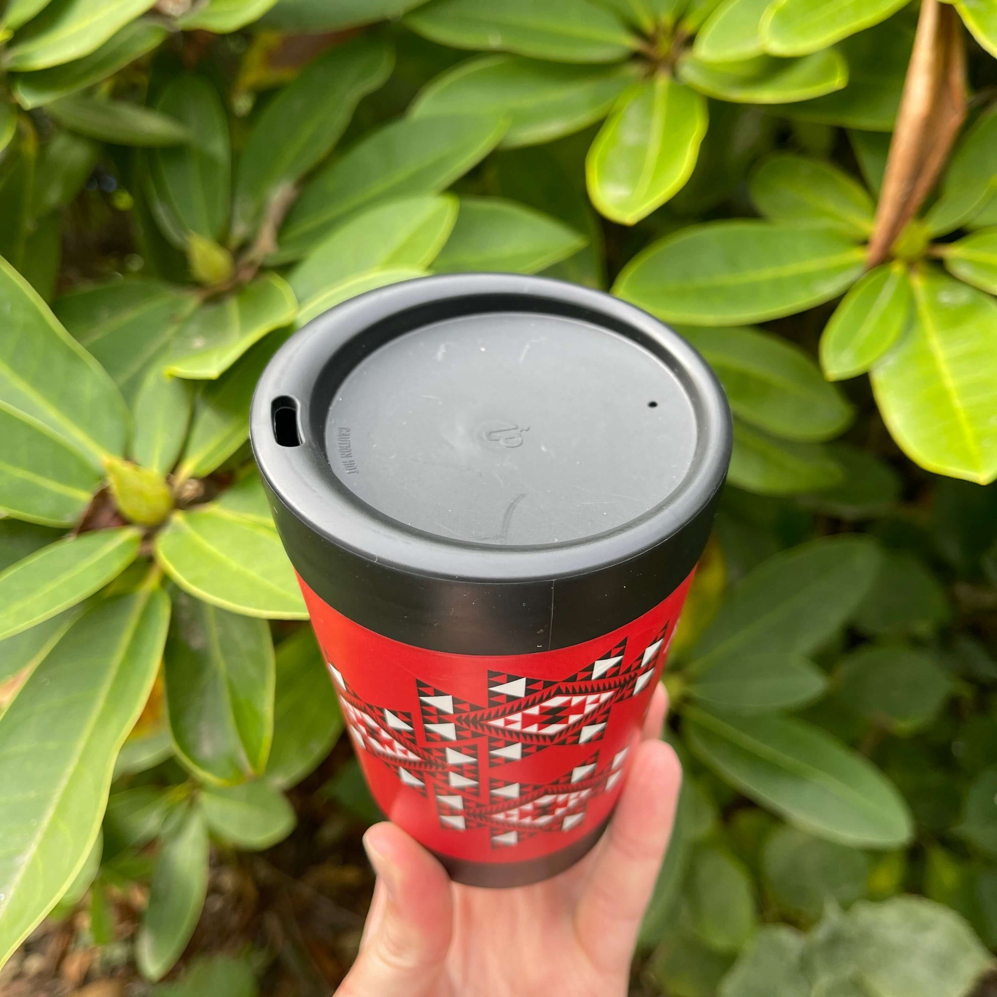 Reusable coffee cup in black with red, white and black wrapped image featuring Maori patterns and showing the lid with sipping hole.