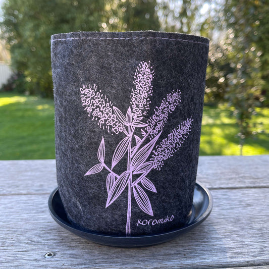 Dark grey felt planter with Koromiko flowers printed on it in purple, sitting in a grey dish.