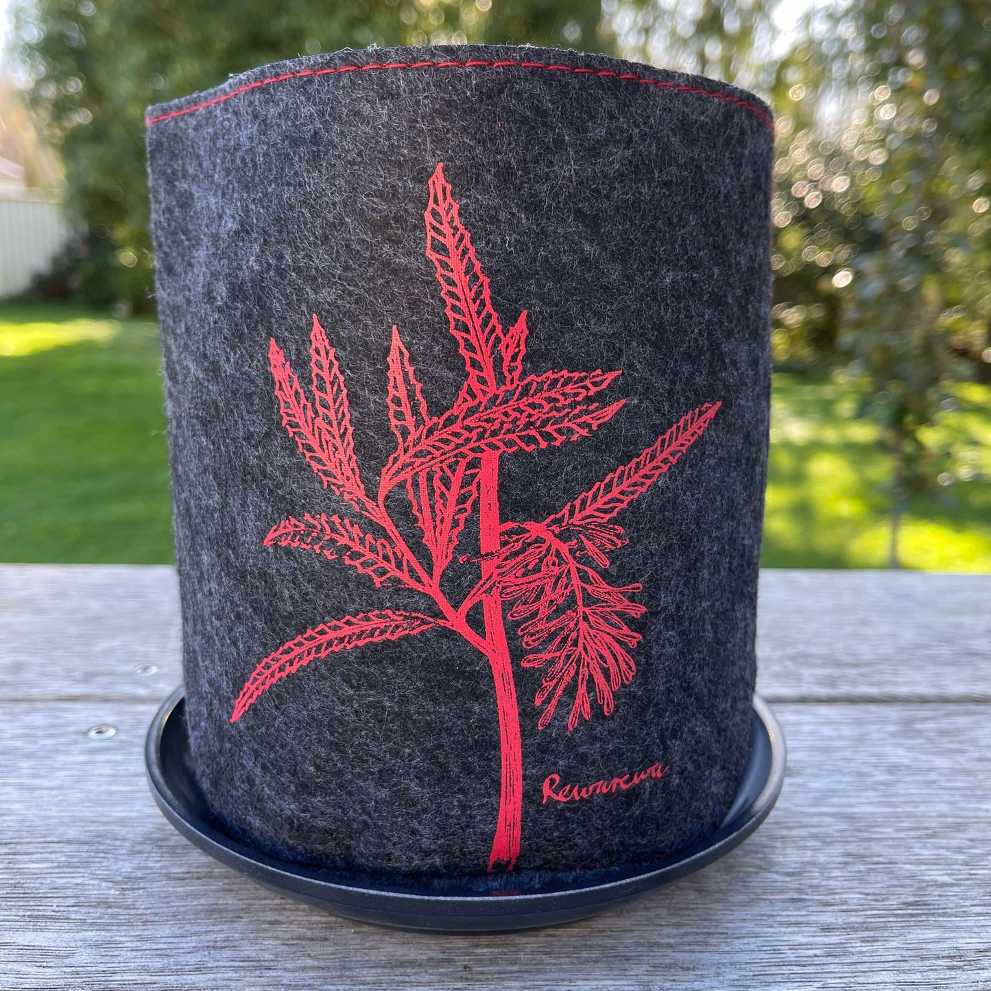 Dark grey felt planter with a rewarewa plant printed on it in red and sitting in a grey dish.