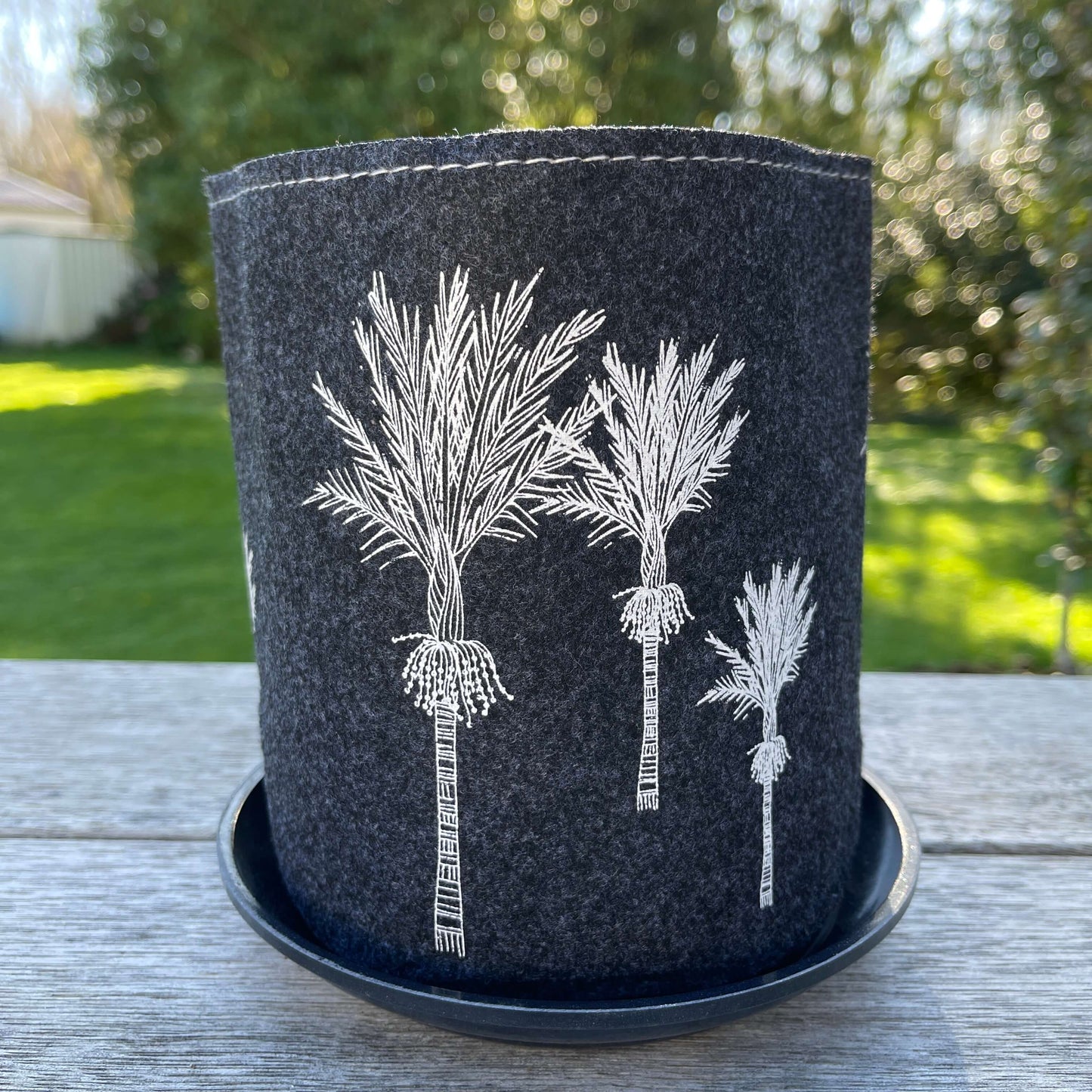 Dark grey felt planter with white Nikau trees printed on it sitting in a grey dish.