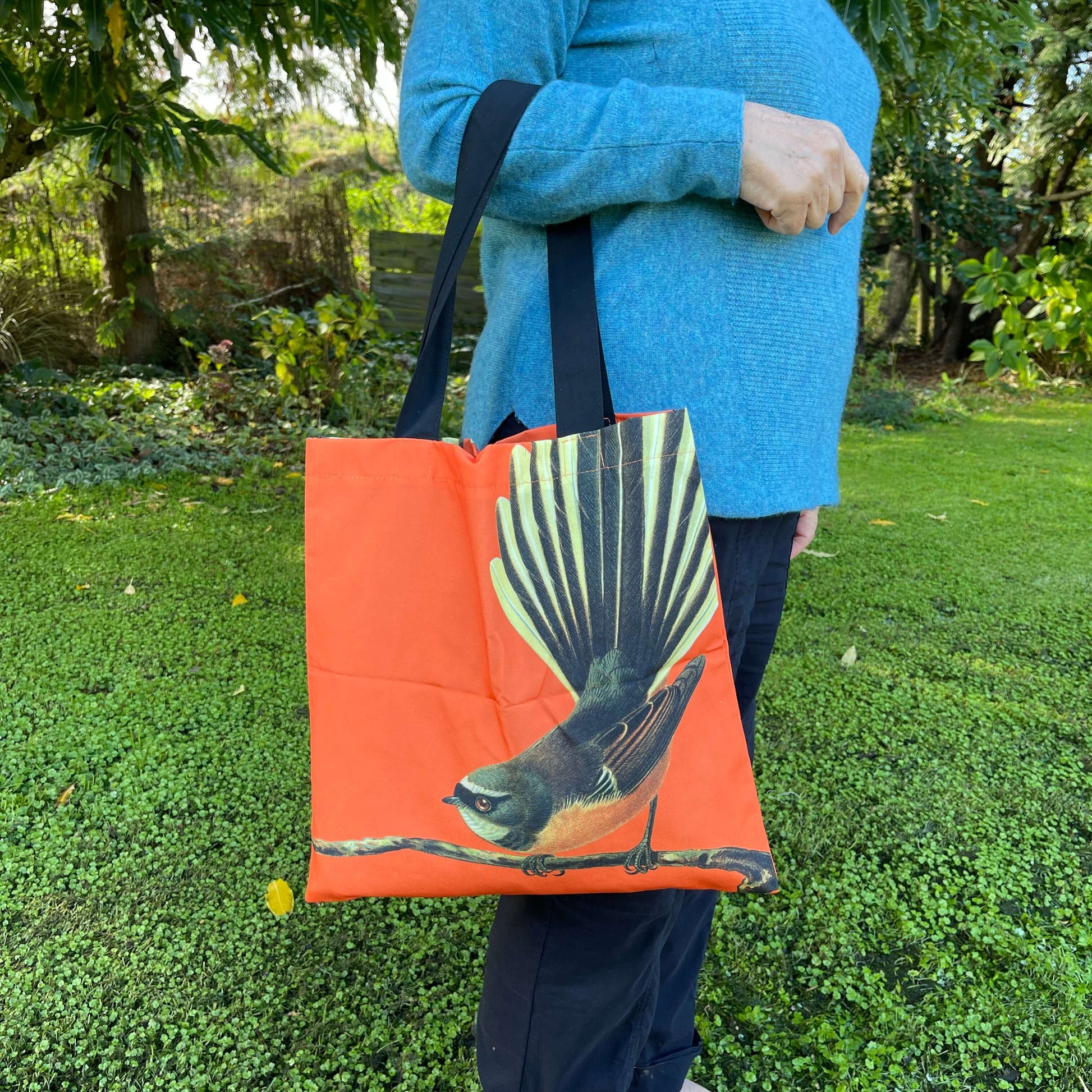 Woman with a tote bag over her arm. The bright orange tote features a fantail bird on a branch.
