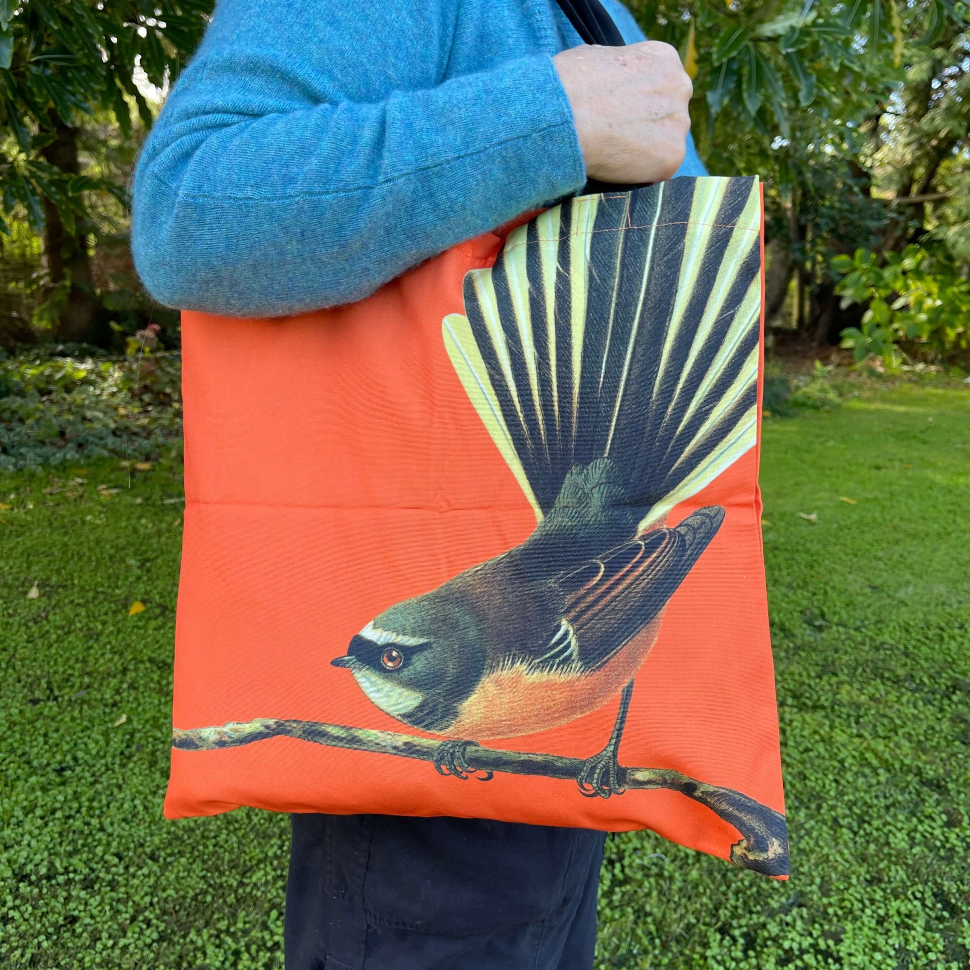 Woman with a tote bag over her arm. The bright orange tote features a fantail bird on a branch.