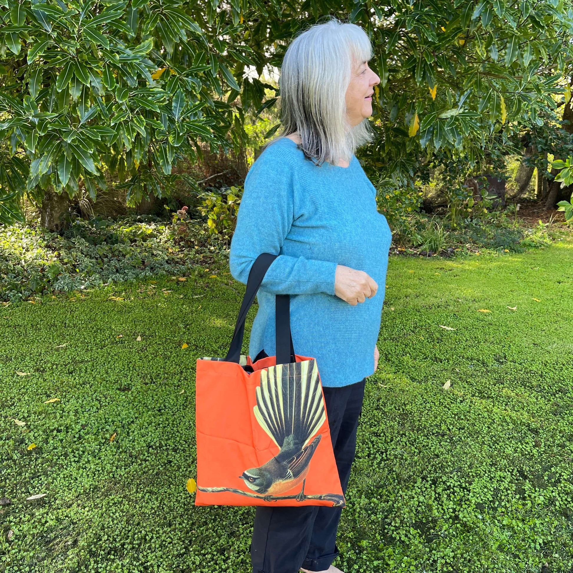 Woman with a tote bag over her arm. The bright orange tote features a fantail bird on a branch.