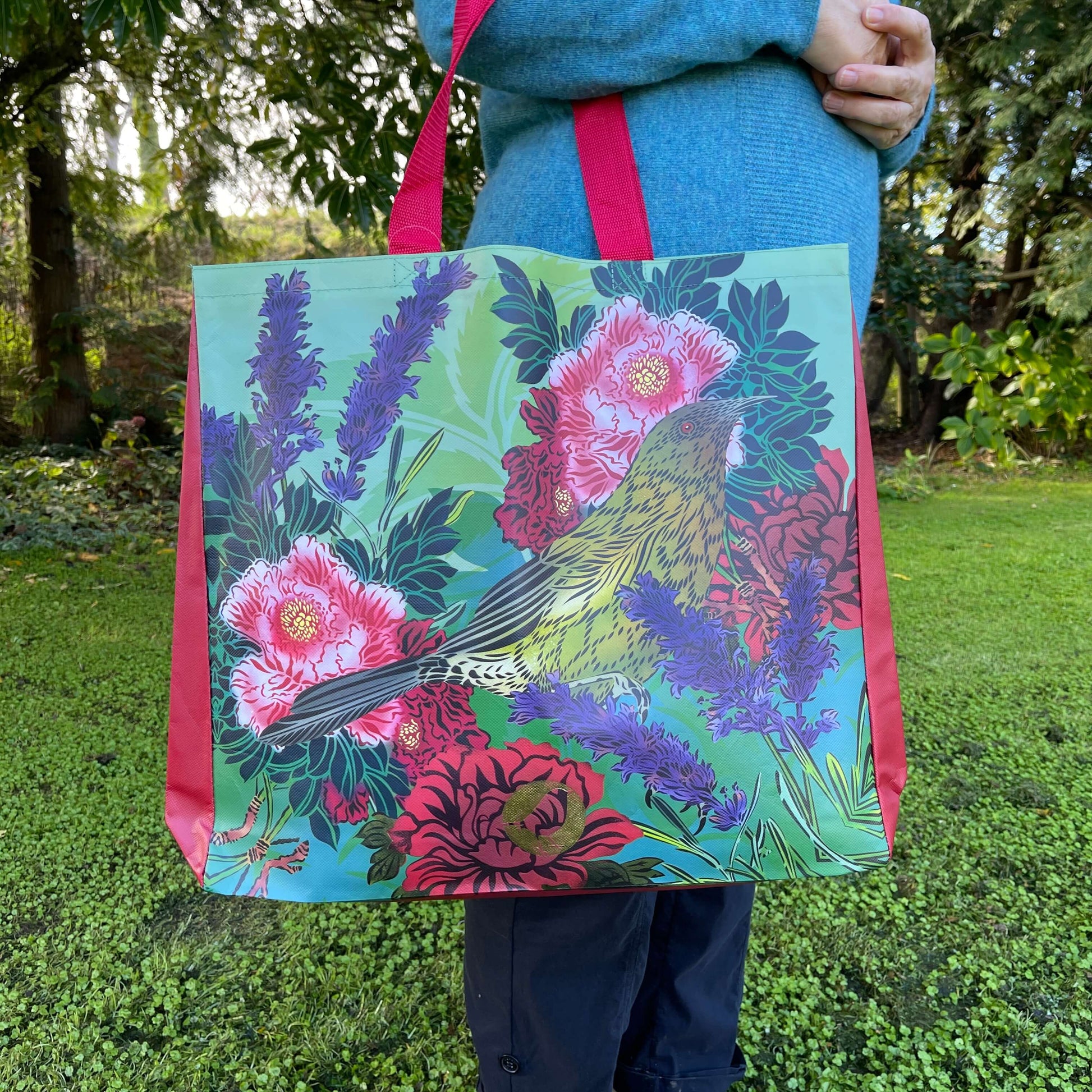 Woman with a tote bag over her arm. The vibrant coloured tote bag features Peony flowers & a bellbird by artist Flox.