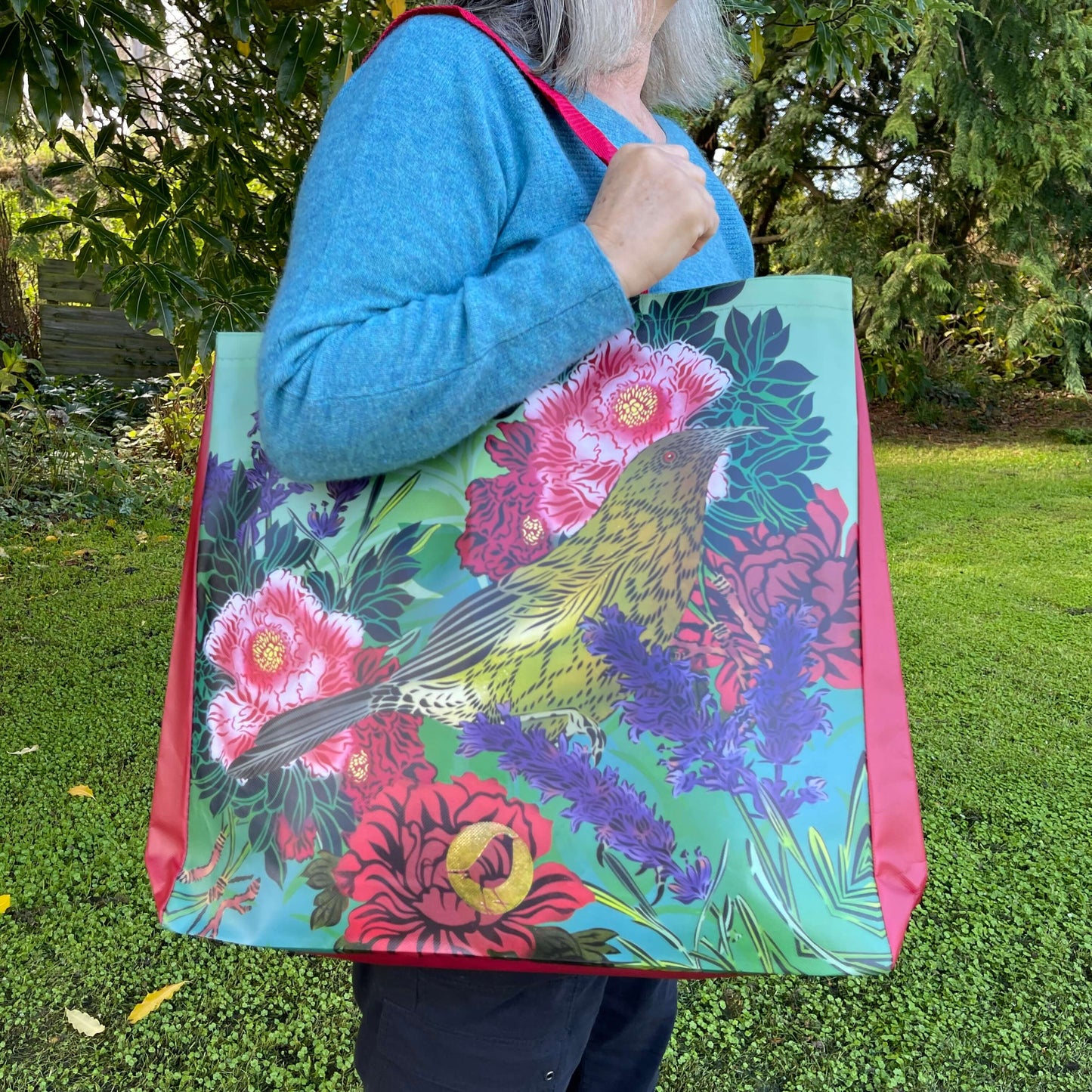 Woman with a tote bag over her arm. The vibrant coloured tote bag features Peony flowers & a bellbird by artist Flox.