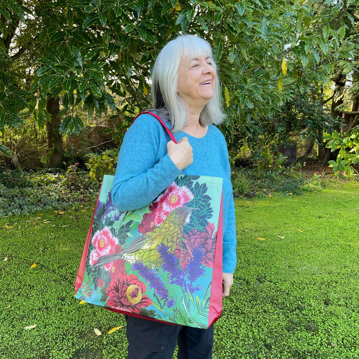Woman with a tote bag over her arm. The vibrant coloured tote bag features Peony flowers & a bellbird by artist Flox.