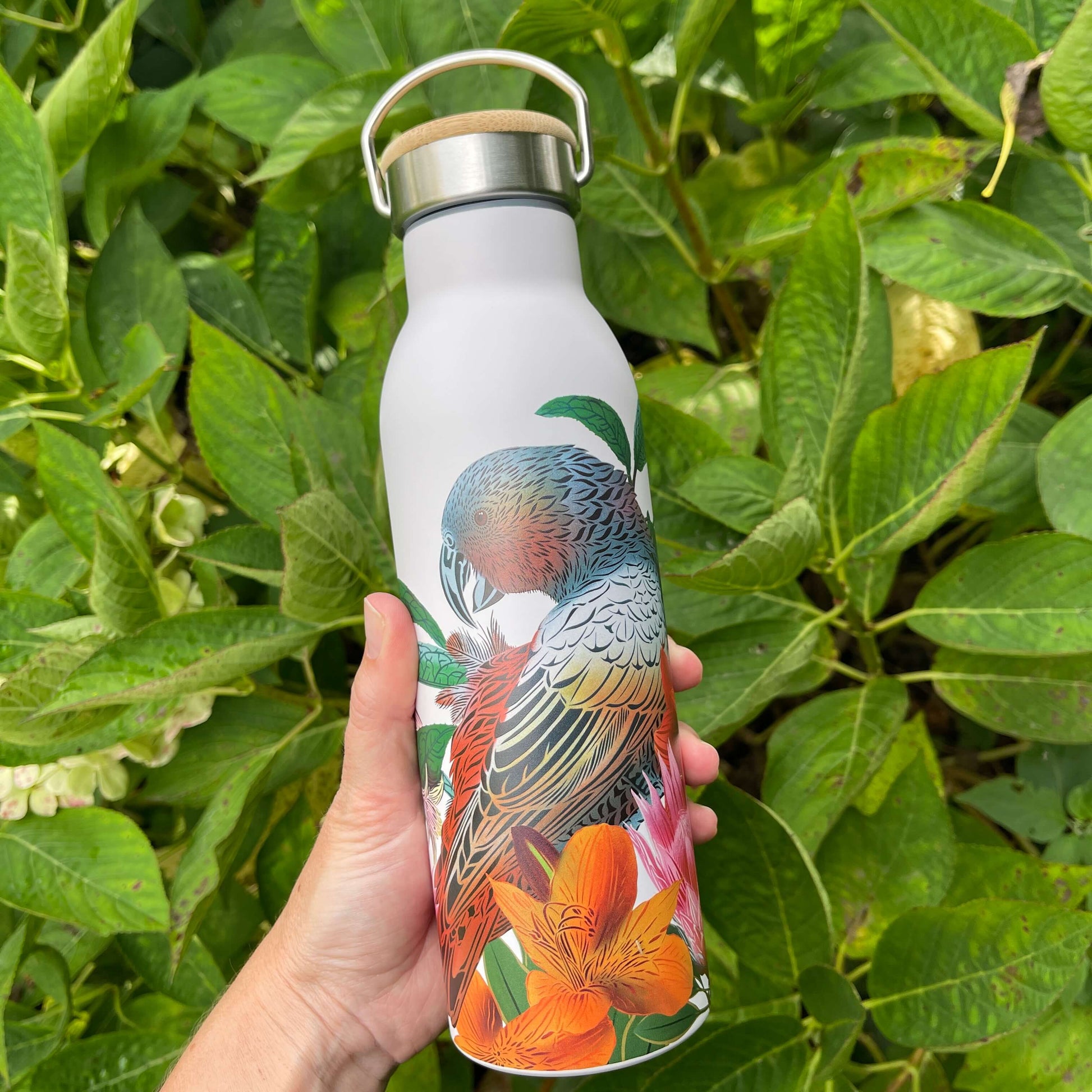 Womens hand holding a white drink bottle featuring artwork of a Kaka bird and flowers. Bottle has a stainless and wooden screw top with handle.