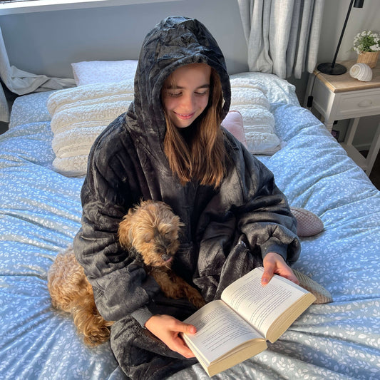 Girl sitting on a bed cuddling a dog and reading a book wearing an oversized grey hoodie.