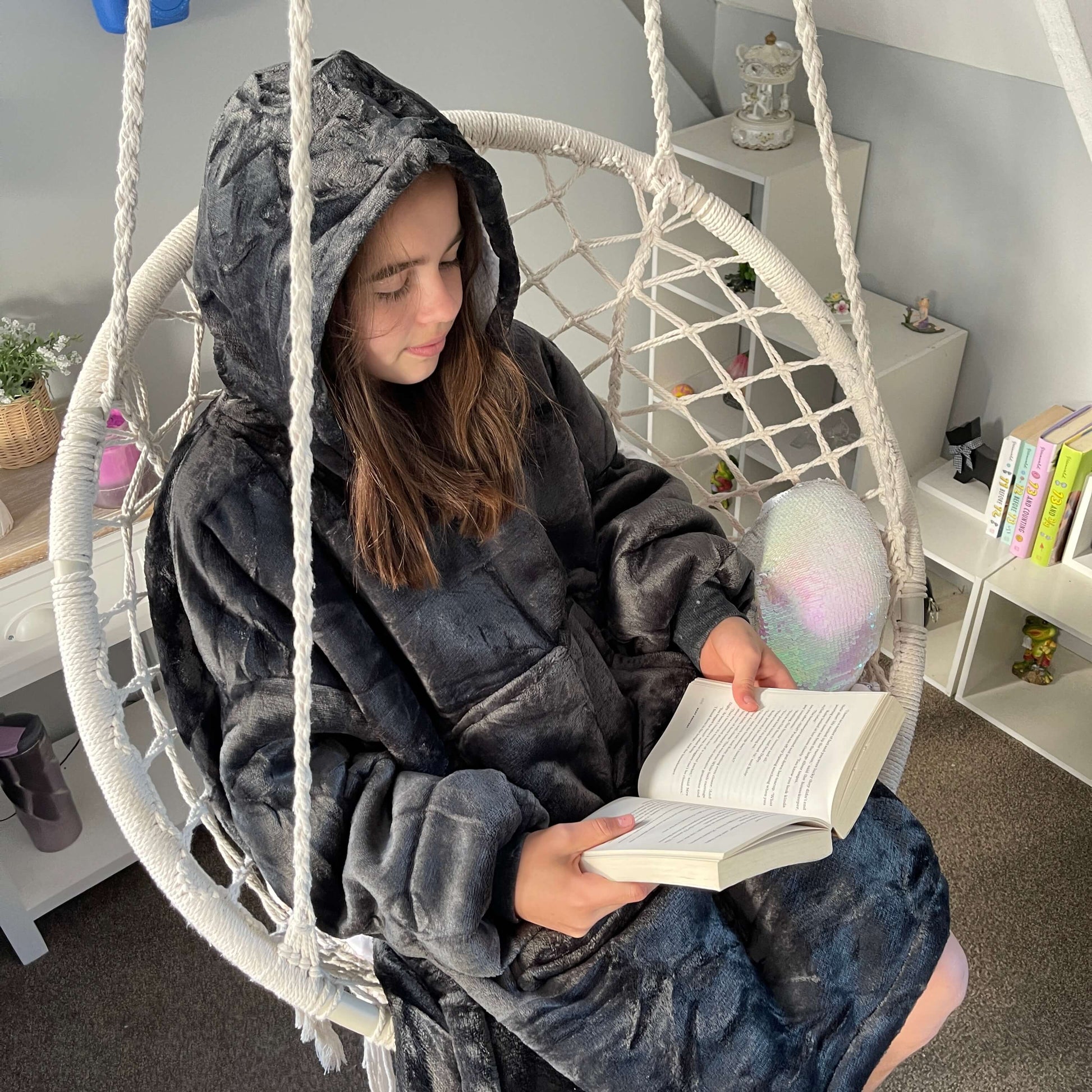 Girl sitting in a swing chair reading a book wearing an oversized grey hoodie.