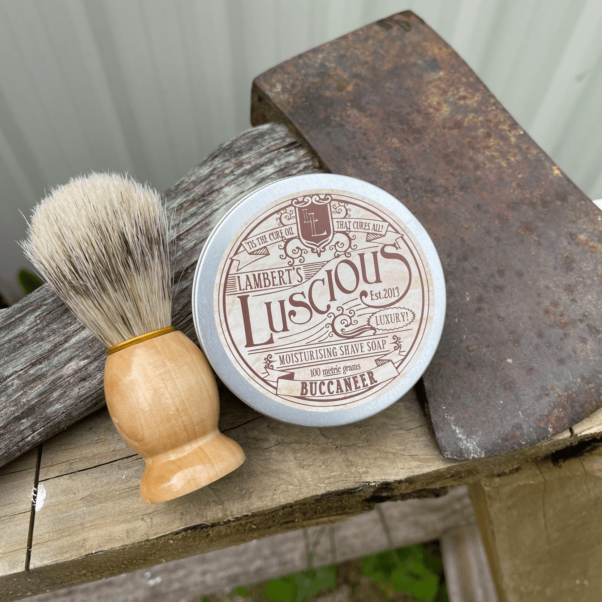 Metal tin of Lamberts Luscious mens shaving soap and a boar bristle shaving brush sitting next to an axe.