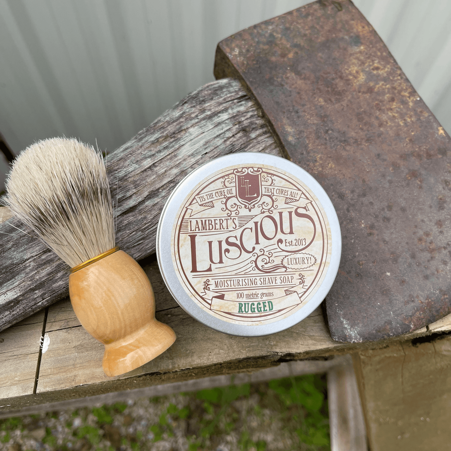 Metal tin of Lamberts Luscious mens shaving soap and a boar bristle shaving brush sitting next to an axe.