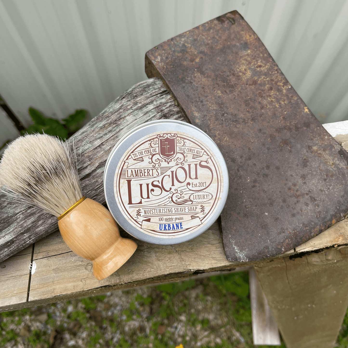  Metal tin of Lamberts Luscious mens shaving soap and a boar bristle shaving brush sitting next to an axe.