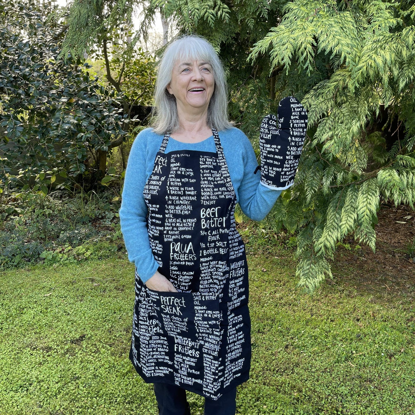 Woman wearing a black apron and mitt set featuring recipes printed on it standing outside.