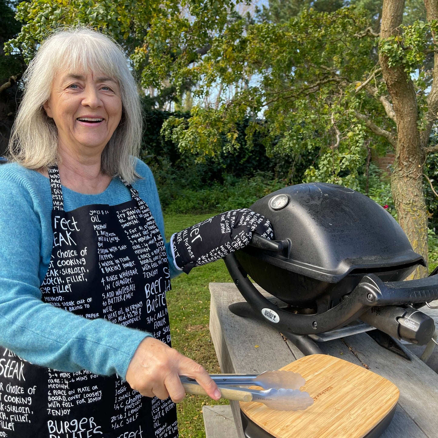 Woman wearing a black apron and mitt set featuring recipes printed on it standing outside at a BBQ.