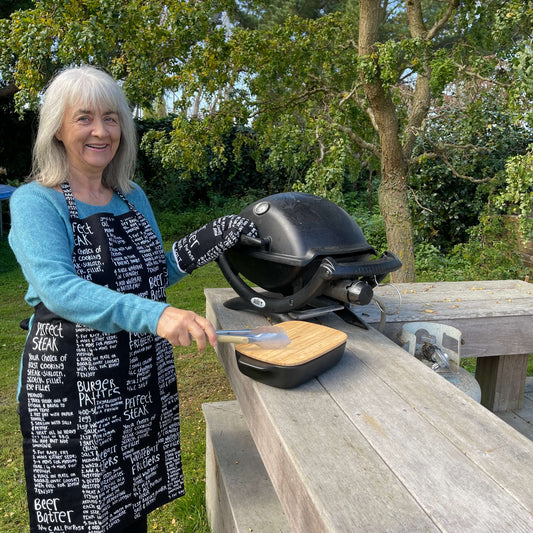 Woman wearing a black apron and mitt set featuring recipes printed on it standing outside at a BBQ.