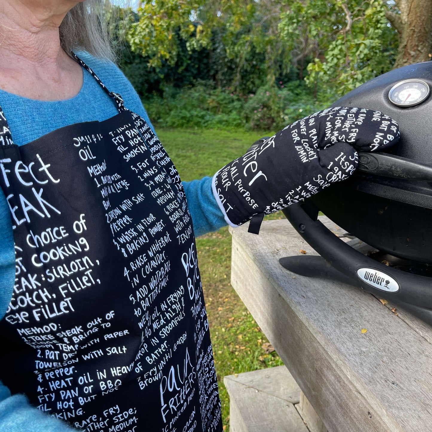 Woman wearing a black apron and mitt set featuring recipes printed on it standing outside at a BBQ.