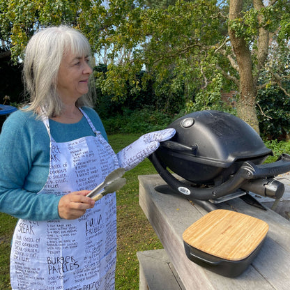 Woman wearing a white apron and mitt set featuring recipes printed on it standing outside at a BBQ.