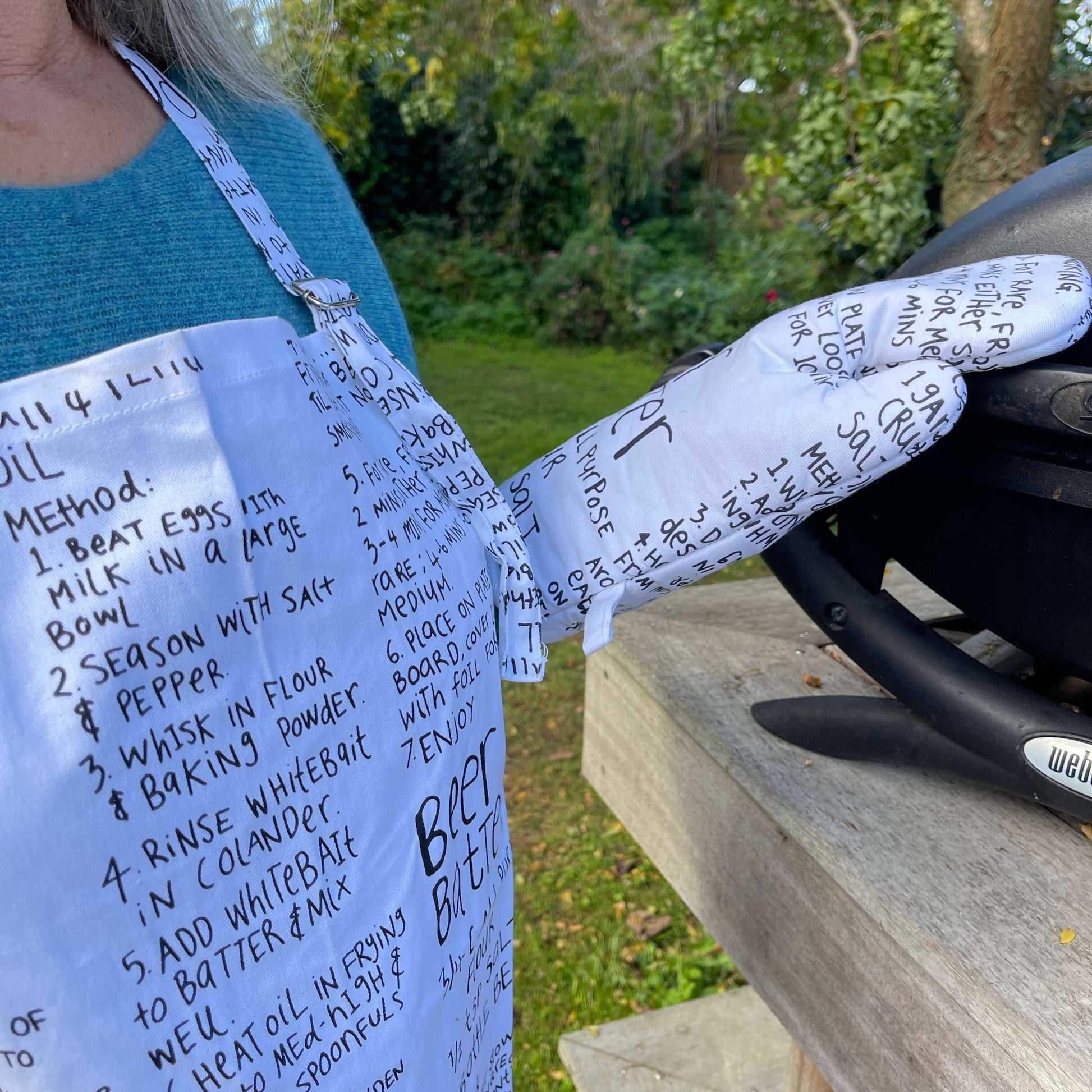 Woman wearing a white apron and mitt set featuring recipes printed on it standing outside at a BBQ.