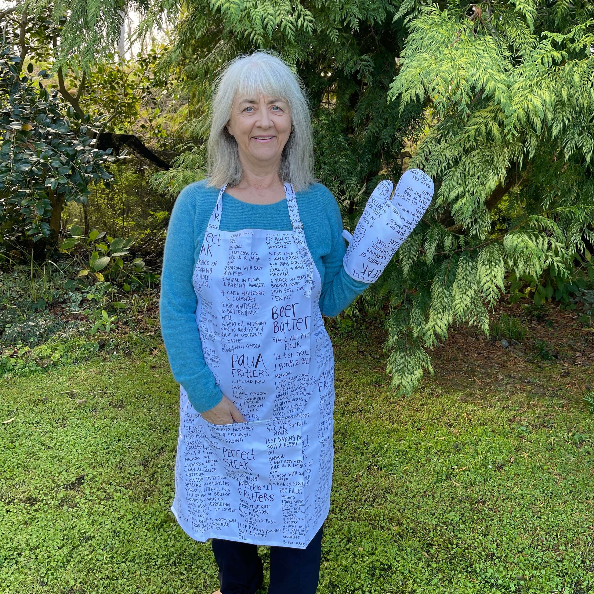 Woman wearing a white apron and mitt set featuring recipes printed on it standing outside.